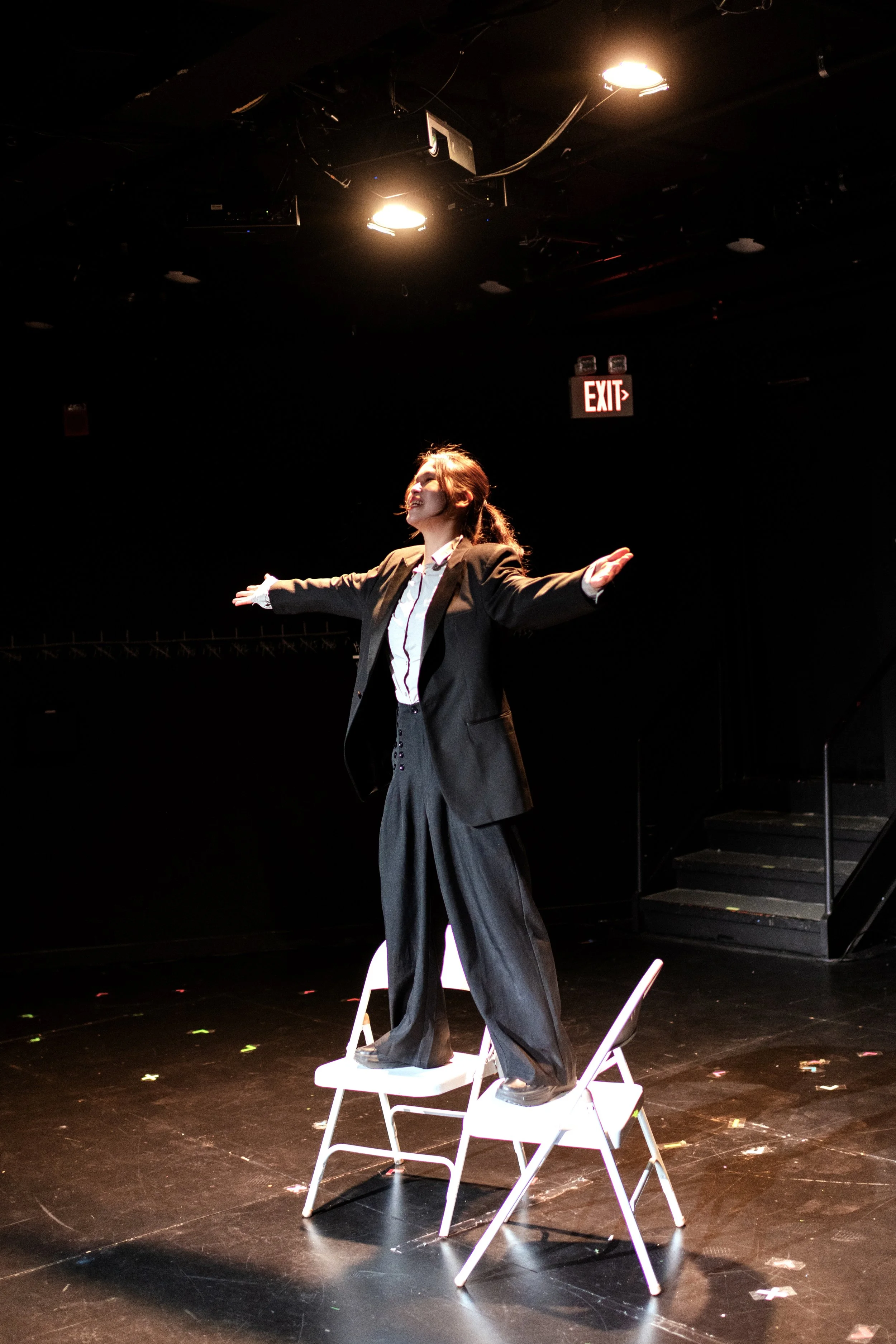 A woman dressed in a suit with arms outstretched standing on two chairs in a dark theater or performance space with stage lights overhead and an exit sign glowing red in the background.