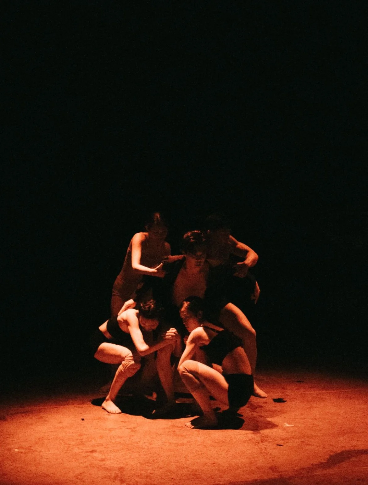 A group of performers engaging in a dance or theatrical performance on a dimly lit stage, with a dark background and focused lighting on the performers.