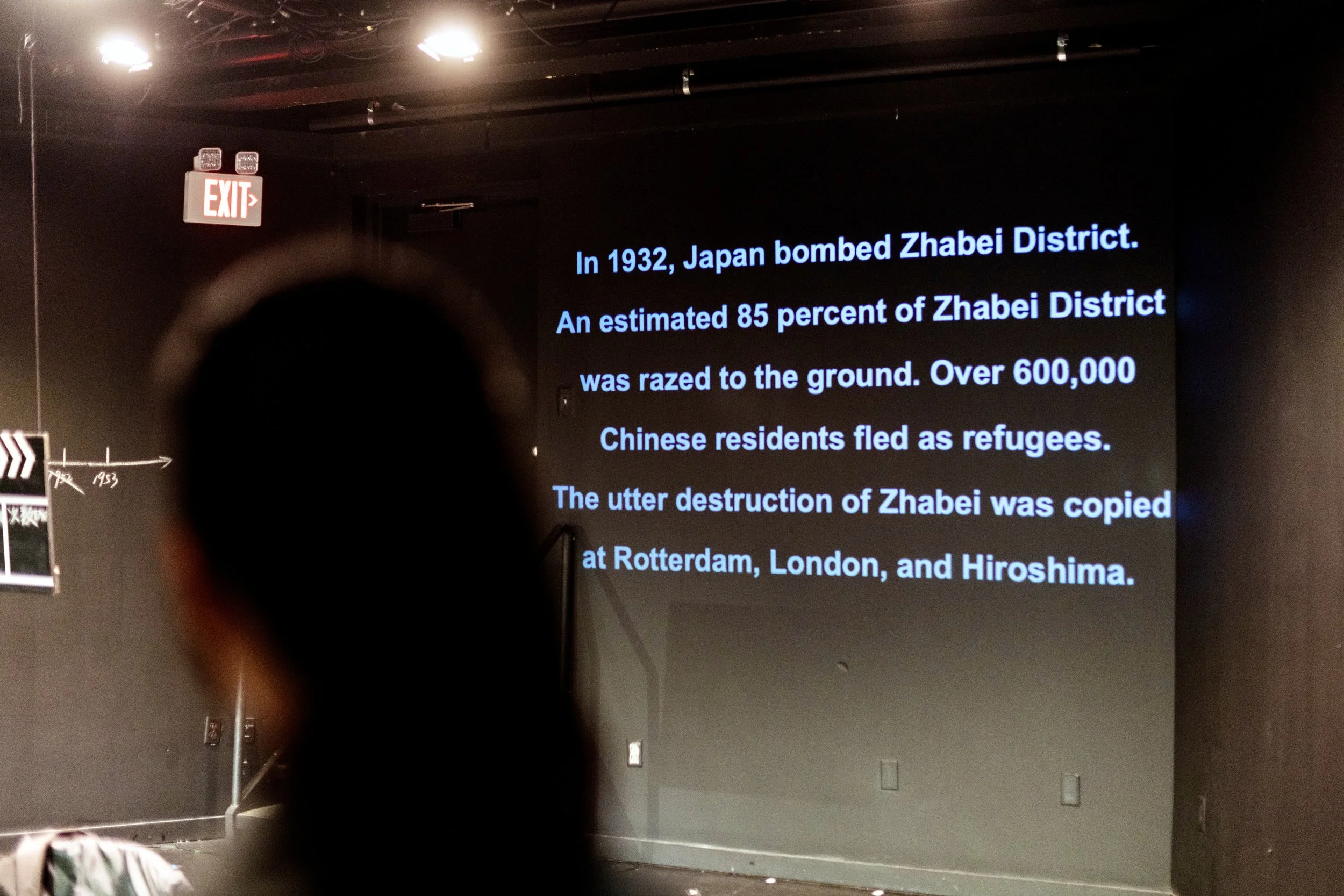 A large screen with text about Japan's bombing of the Zhabei District in 1932, including details about refugees and destruction, in a dark room with an exit sign overhead.