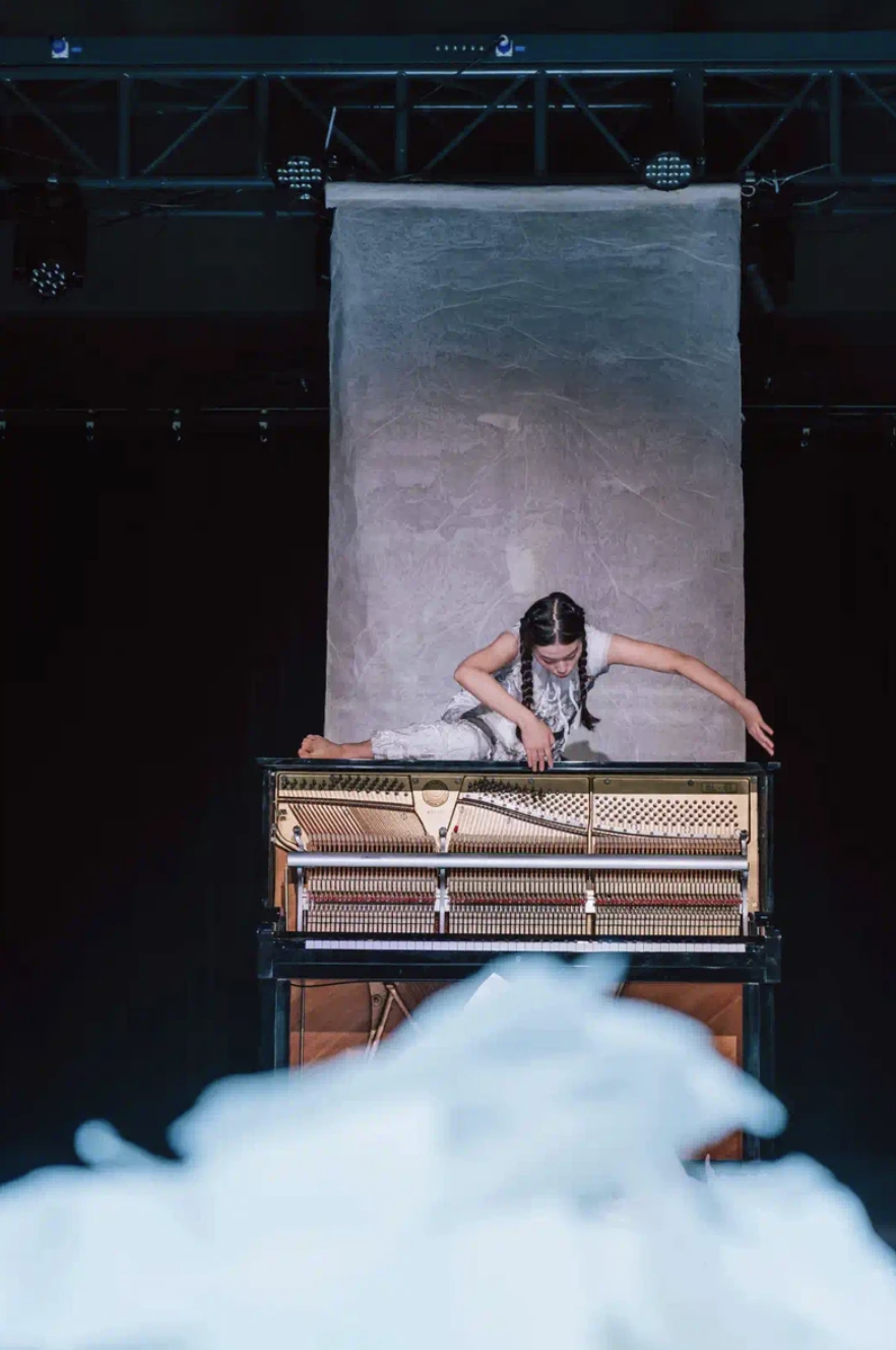 A young girl with braided hair in a white dress on an open grand piano, leaning over it on stage with a gray backdrop, stage lights, and smoke at the foreground.