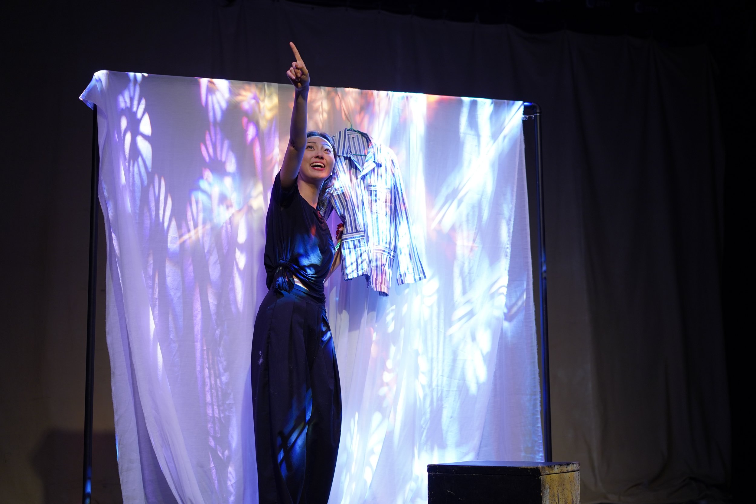 A woman standing on stage, pointing upward with her right hand, smiling, next to a hanging striped shirt, with colorful light patterns projected on a white curtain behind her.