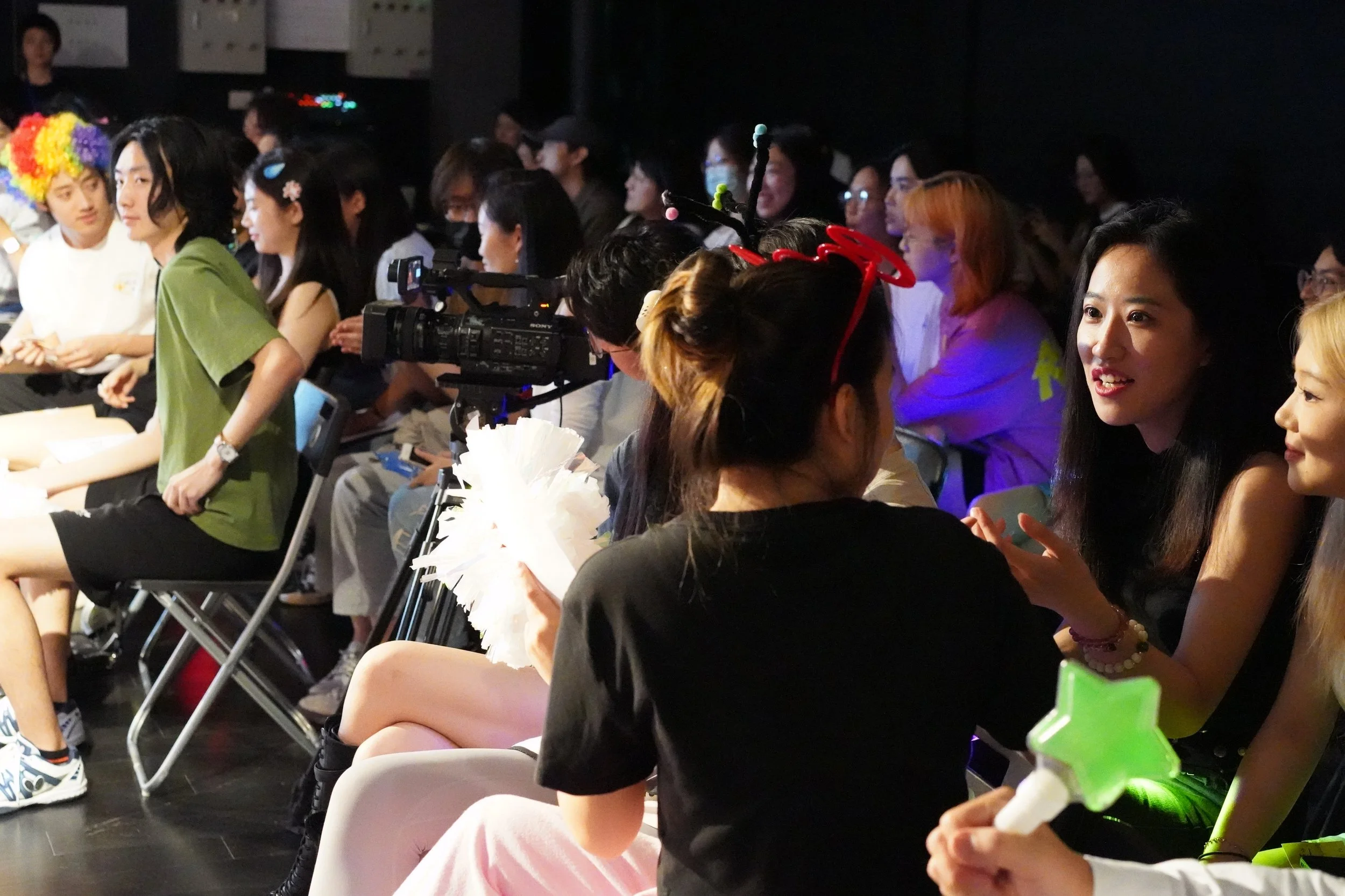 A group of people seated in rows, attending an event, with some wearing colorful accessories and costumes.