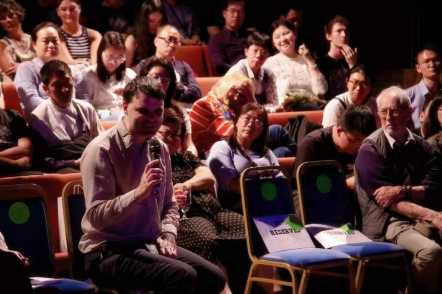 Audience sitting in theater seats, some taking notes or holding microphones, attending a presentation or event.