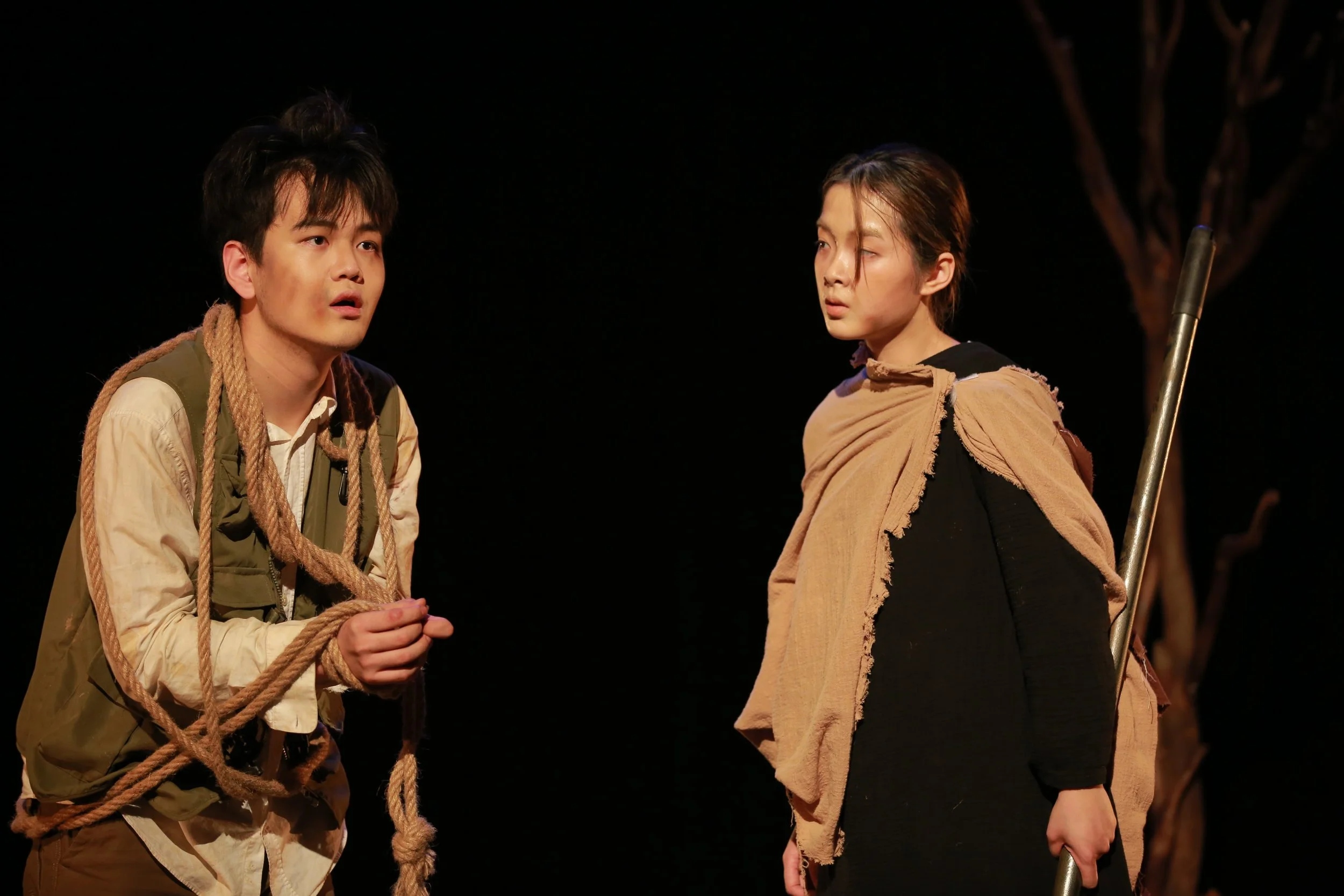 Two actors on stage, one with short dark hair and the other with tied-back hair, engaged in a scene. The actor on the left is holding a rope, while the actor on the right is standing with a stick. The background is dark.
