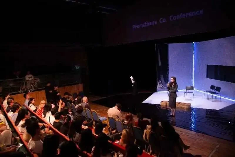 A woman in formal attire speaking on stage at a conference with a seated audience and a projected sign reading 'Prometheus Co. Conference' above.