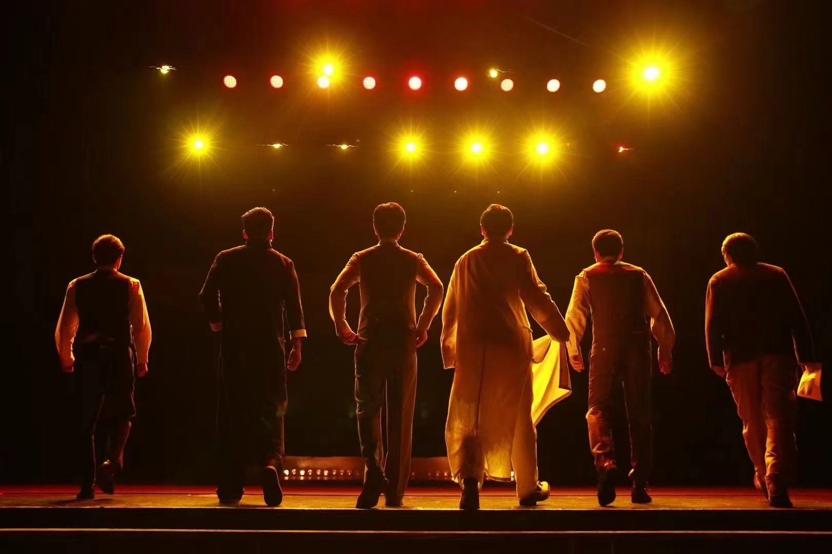 Six performers in formal attire standing on stage with back to audience, facing bright stage lights at night