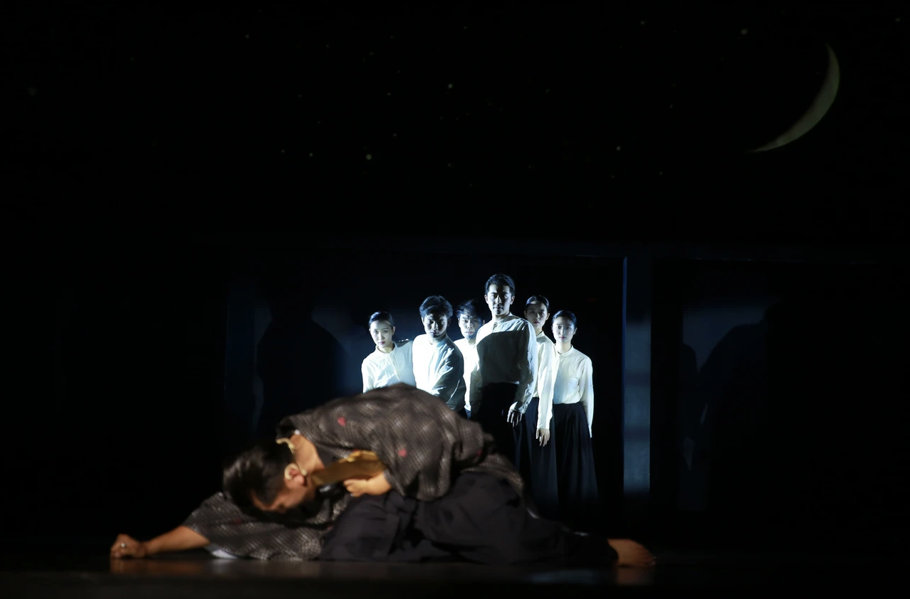 A performer bowing on stage with a background projection of six other performers standing in a line, illuminated against a dark backdrop with a crescent moon and stars.