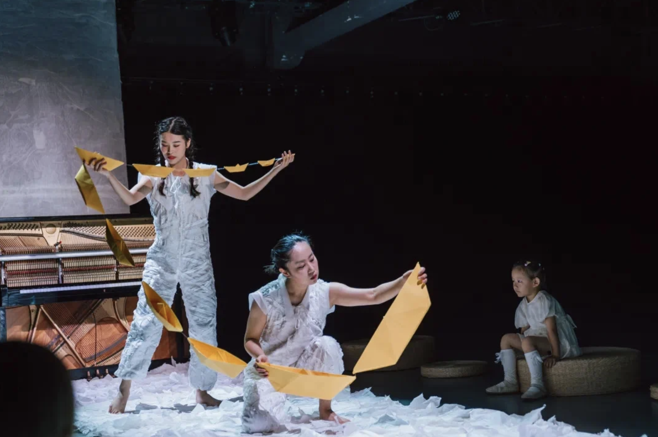 A group of young girls performing a dance on stage, holding yellow paper umbrellas, with a piano in the background and a dark stage environment.