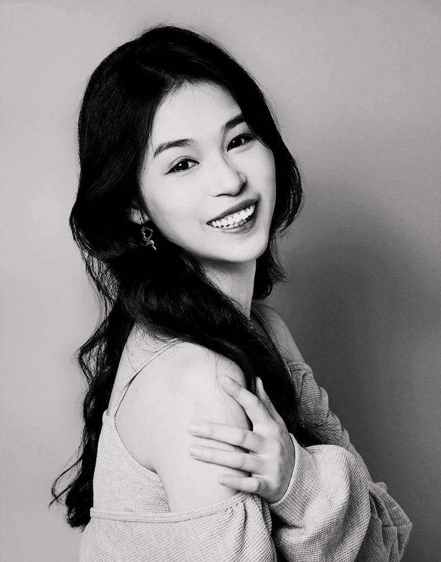 Black and white portrait of a young woman smiling, with long dark hair, wearing earrings and a sweater that falls off one shoulder, standing against a plain background.