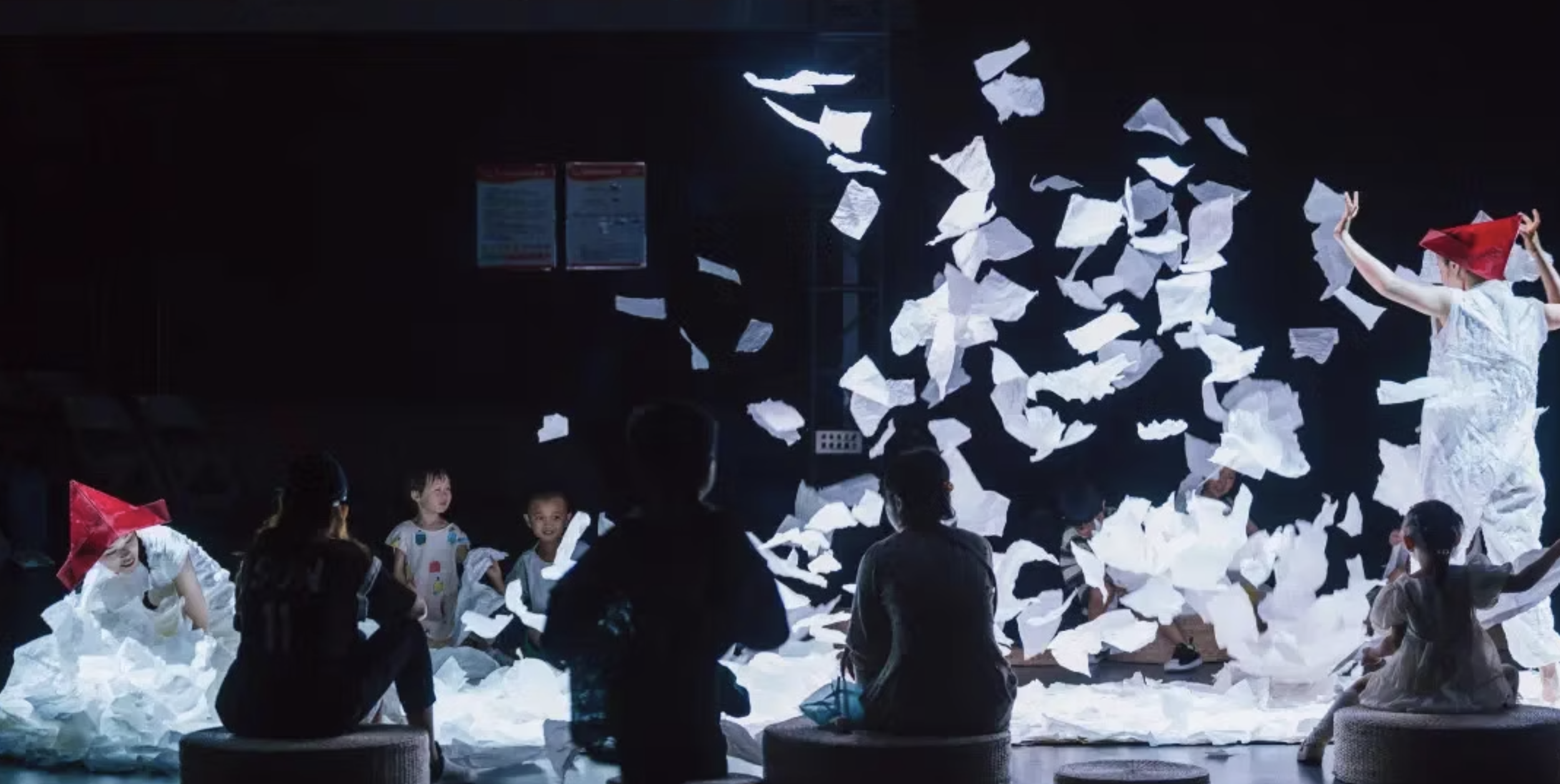 Children and a performer on stage surrounded by falling white papers, with some children wearing paper hats.