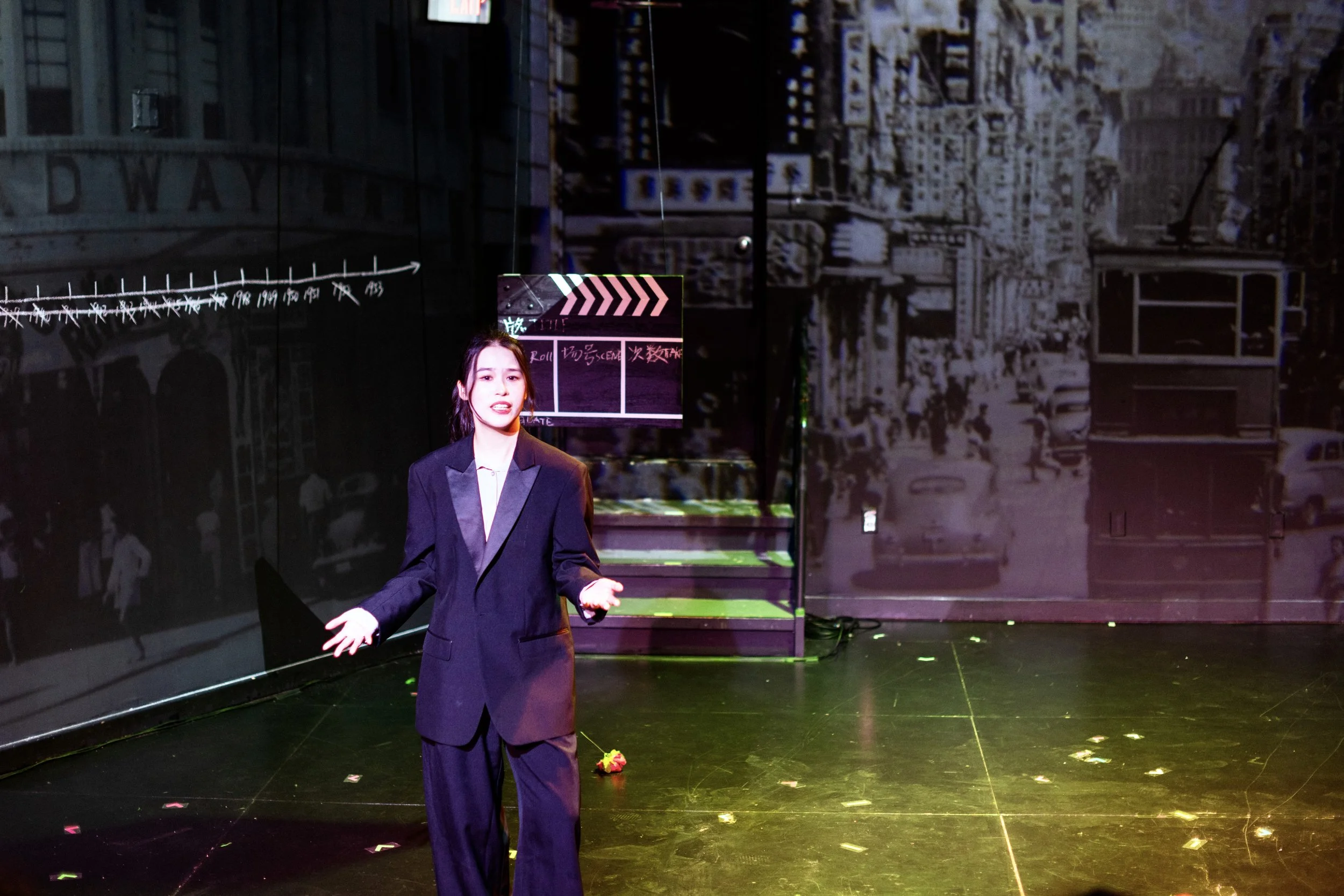 A woman with long dark hair, dressed in a black blazer and white shirt, standing on a stage with a cityscape background and a director's clapboard sign behind her. She appears to be speaking or performing.
