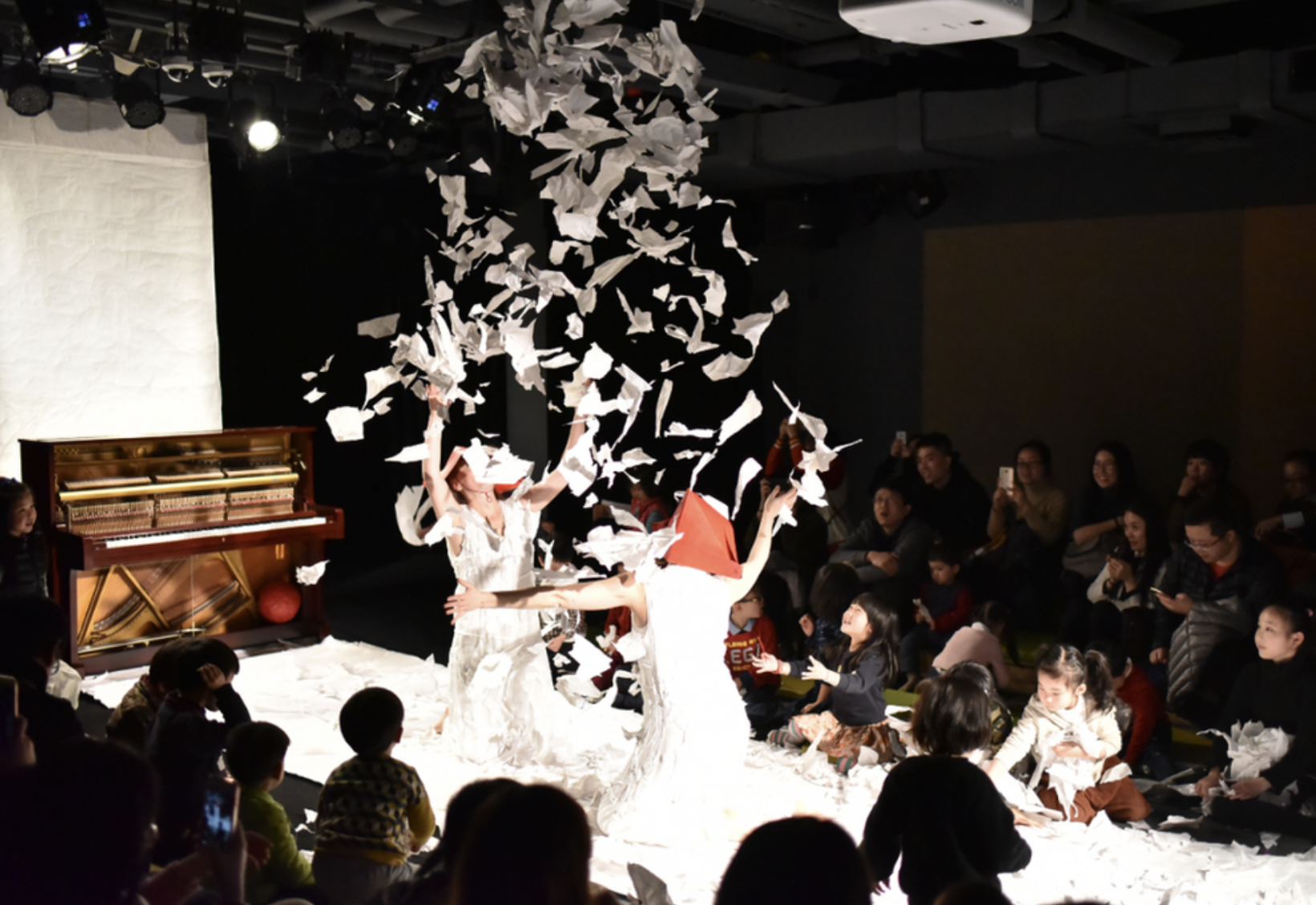 A stage performance with two women in white dresses surrounded by falling paper, with an audience watching and taking photos.