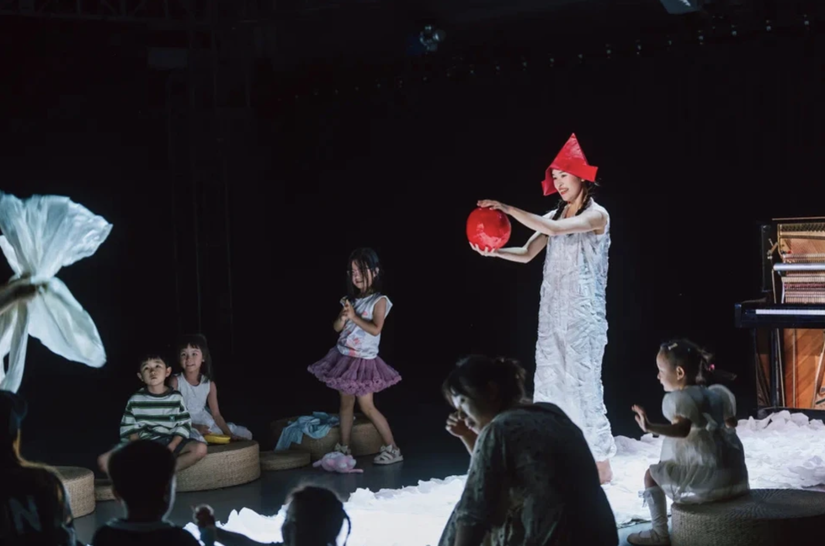 A woman in a white dress wearing a red paper hat, holding a red paper lantern, performing on stage with children around her. Some children are sitting on stools, others are standing or kneeling, engaging with the performance. The stage has a black background with stage lighting, and there is a piano on the right side.