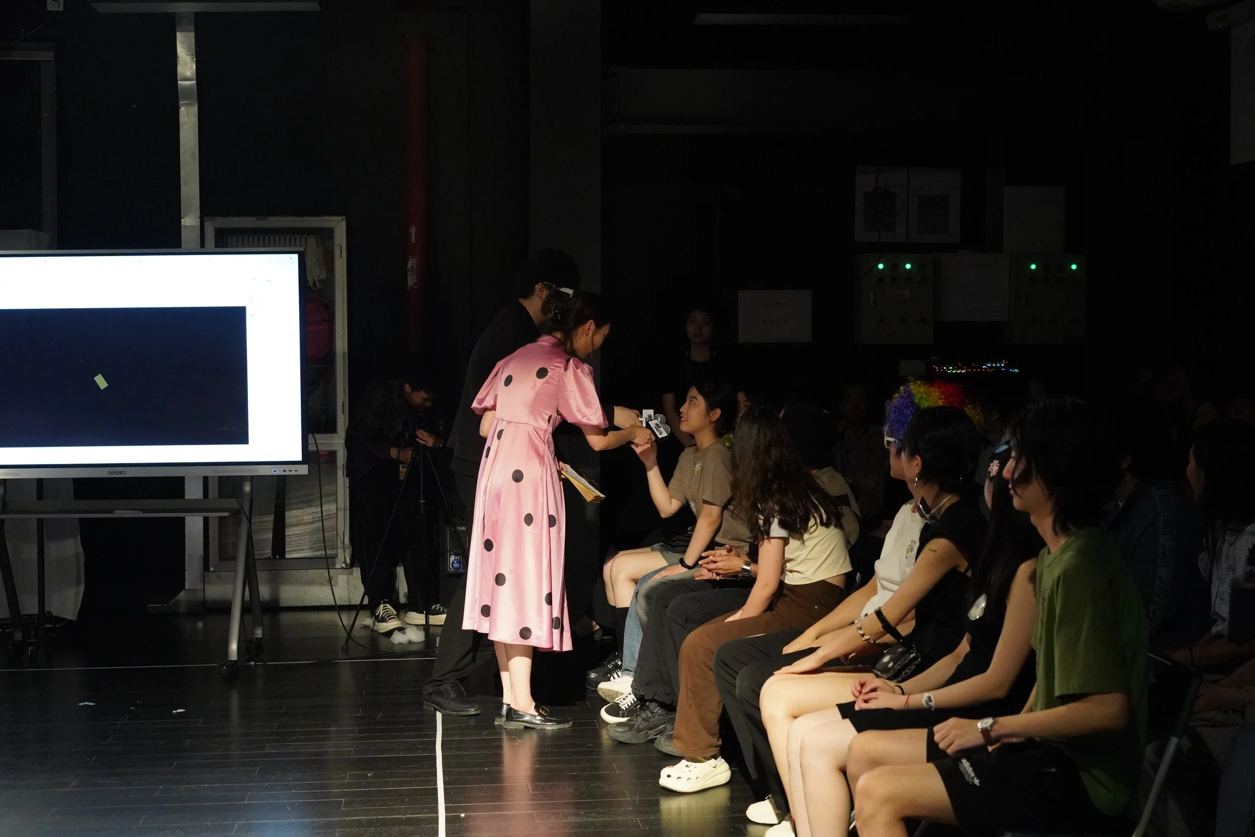 A woman in a pink dress with black polka dots is handing a sash or certificate to a seated young woman during an award or recognition ceremony in a dark room, with an audience and a large screen nearby.