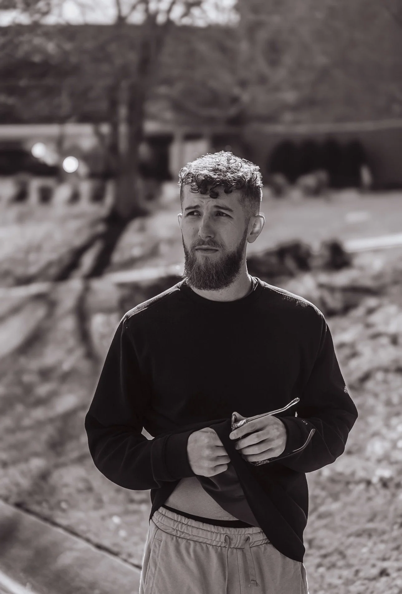 A young man with a beard and curly hair standing outdoors, holding sunglasses, wearing a black long sleeve shirt and beige shorts, with trees and a park bench in the background.