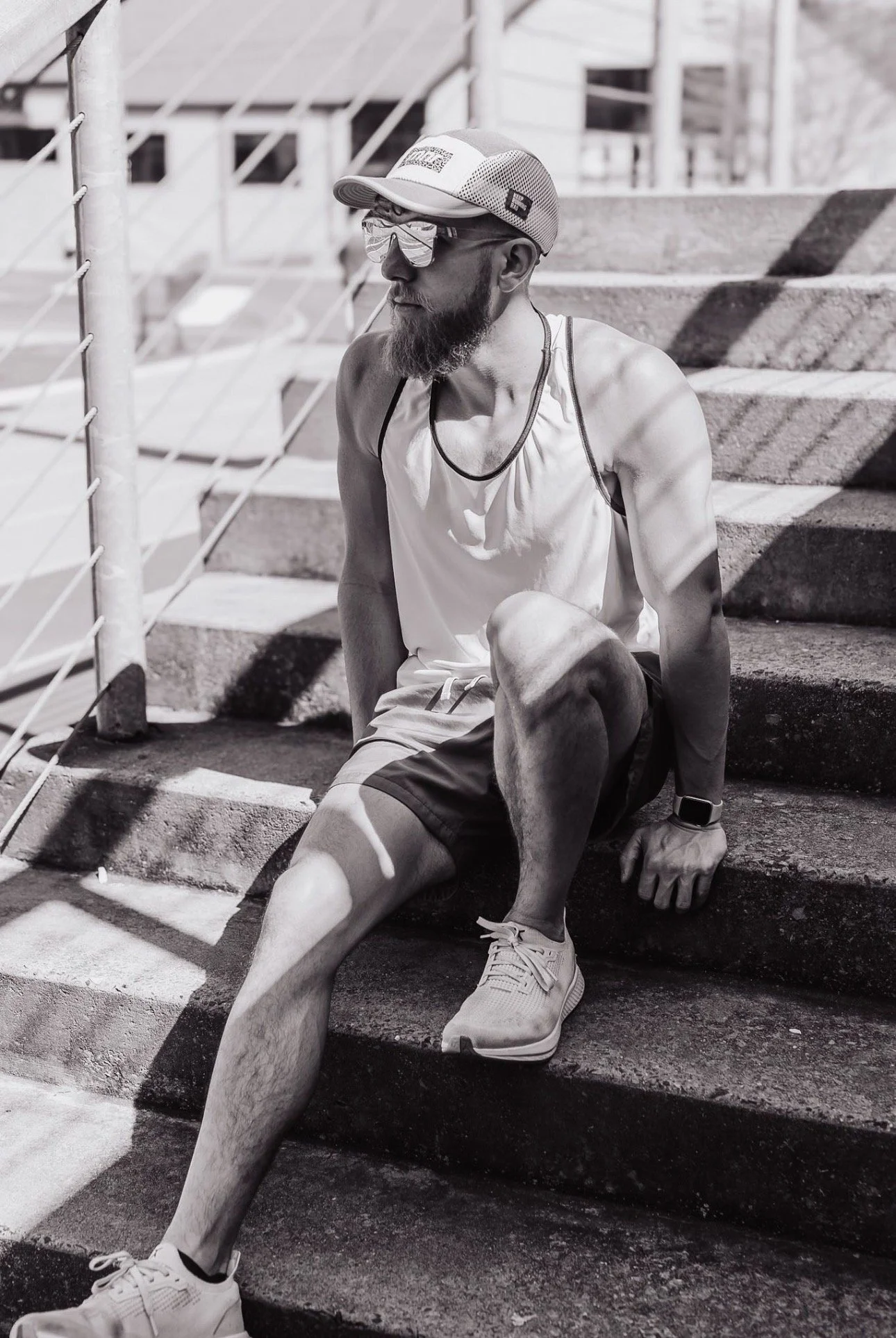 A man with a beard and sunglasses sitting on outdoor concrete stairs, wearing a cap, athletic tank top, shorts, and sneakers, with shadows cast on him from nearby fencing.