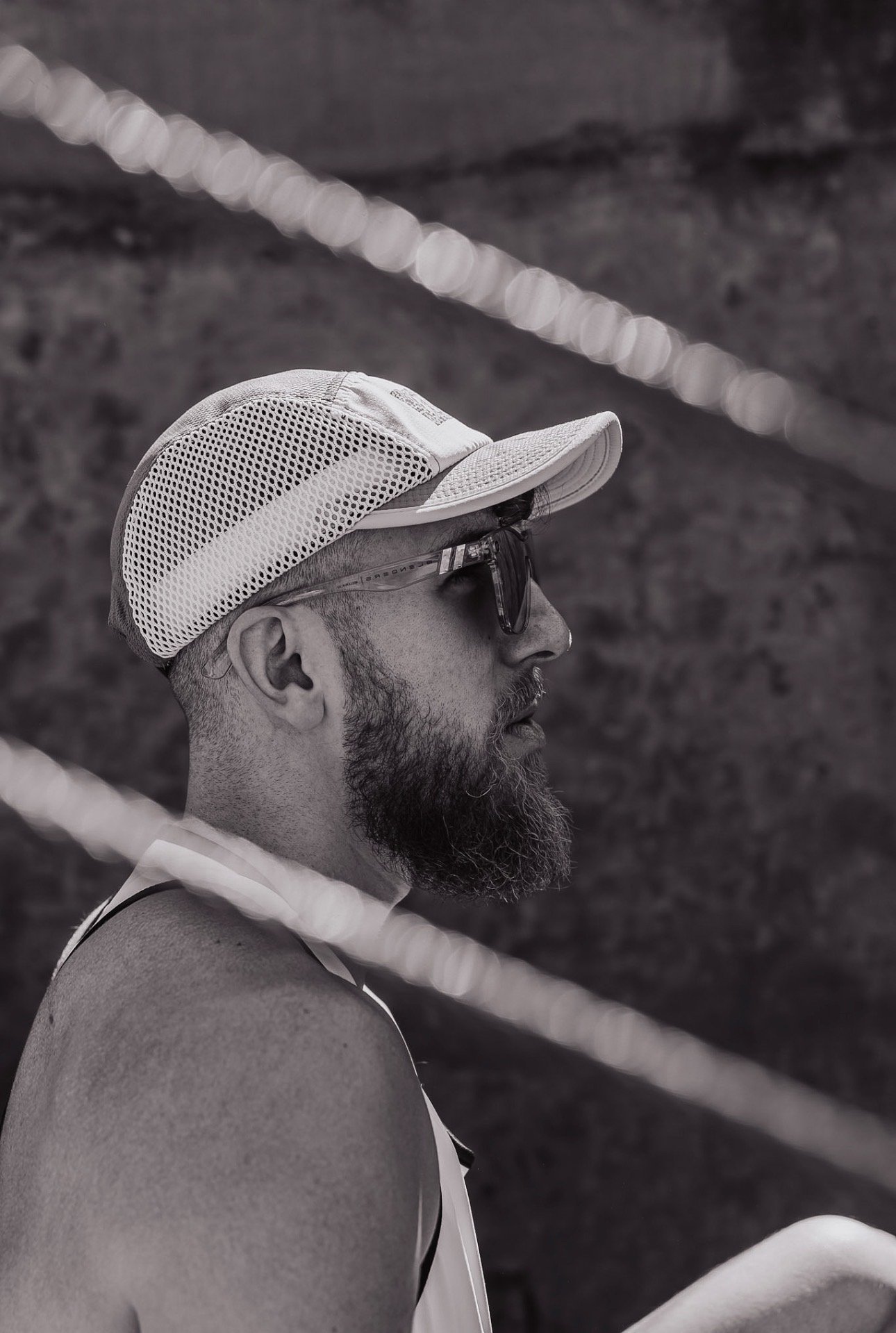 A side profile of a man with a beard wearing sunglasses, a white baseball cap, and a sleeveless athletic shirt, outdoors with blurred background and ropes.