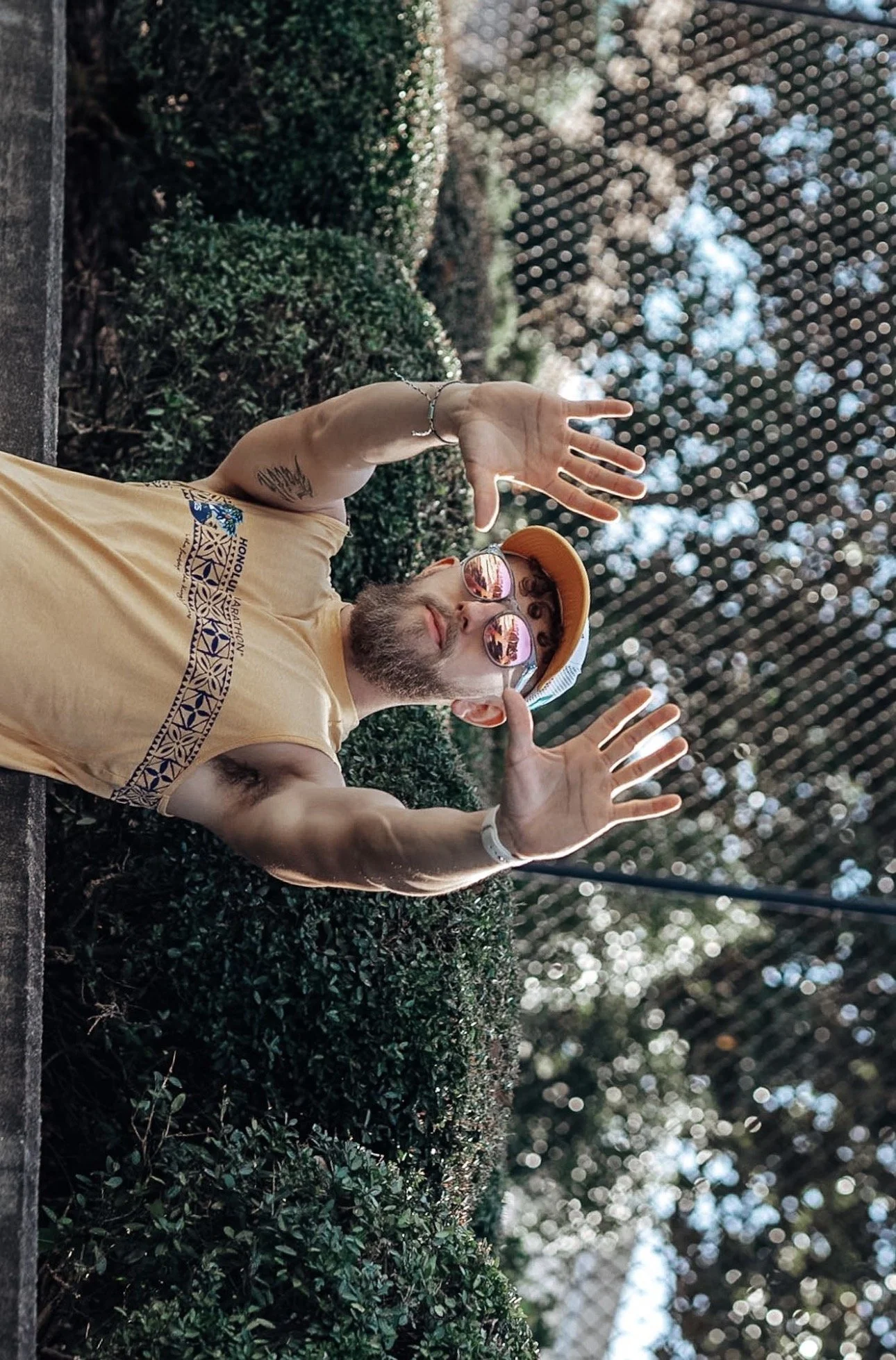 A man wearing sunglasses, a yellow cap, and a sleeveless shirt standing outdoors with his hands raised, against a background of greenery and trees, with sunlight filtering through the leaves.