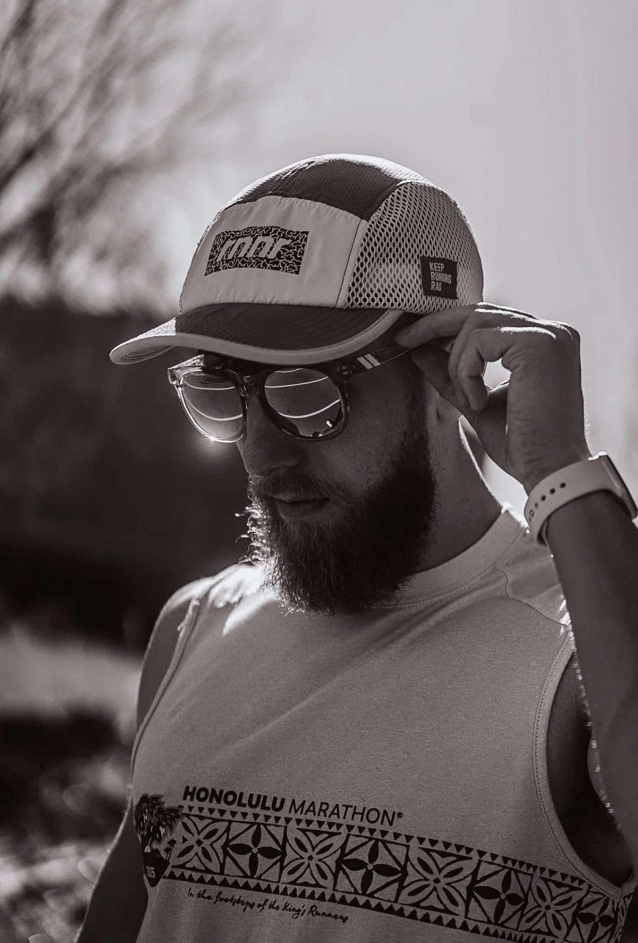 A man with a beard and glasses adjusting a baseball cap, wearing a tank top with 'Honolulu Marathon' written on it, outdoors in bright sunlight.