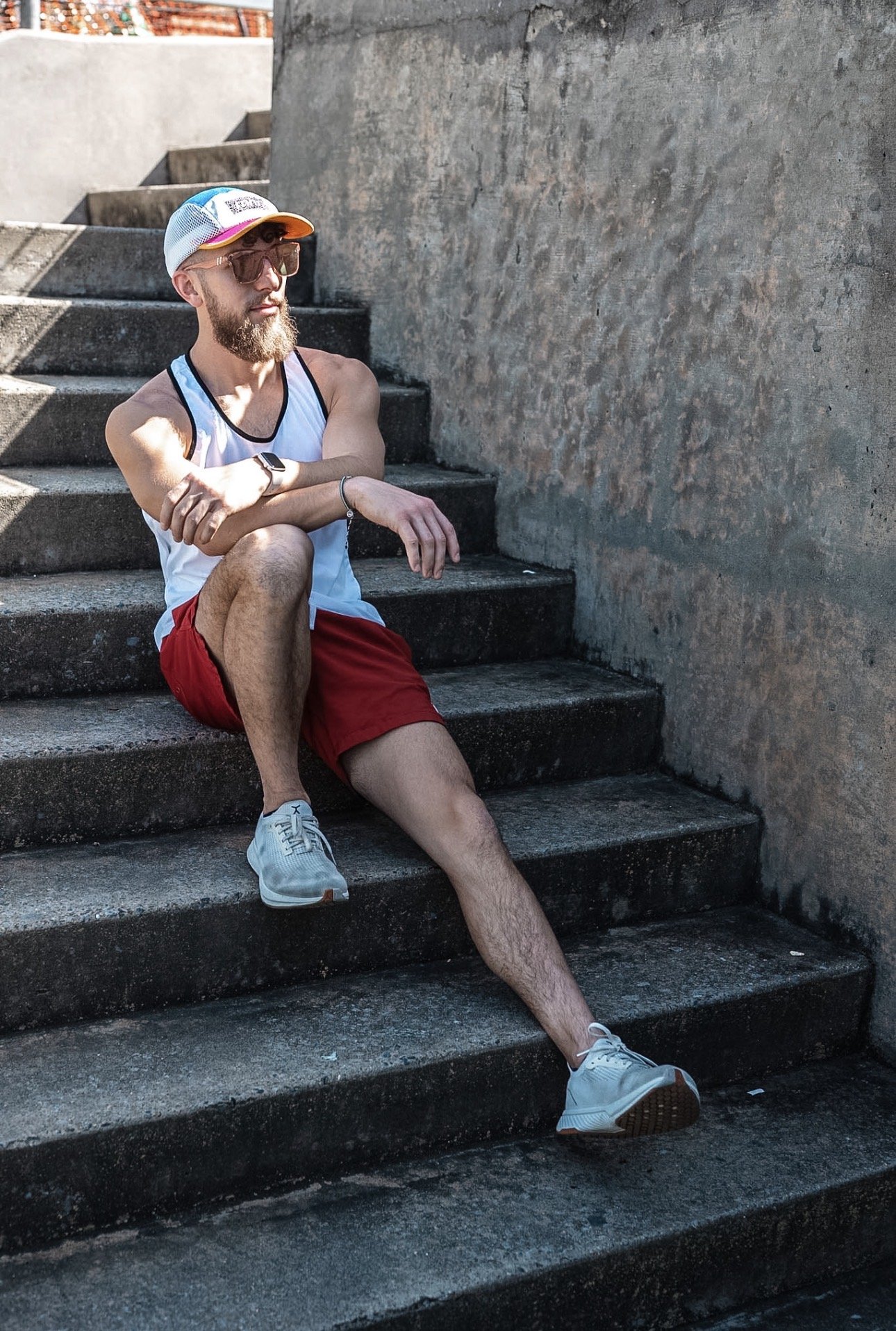 A man with a beard, sunglasses, a colorful cap, a white tank top, red shorts, and white sneakers is sitting on outdoor concrete stairs near a plain, textured wall. He has his arms crossed and one leg stretched out, looking to the side.