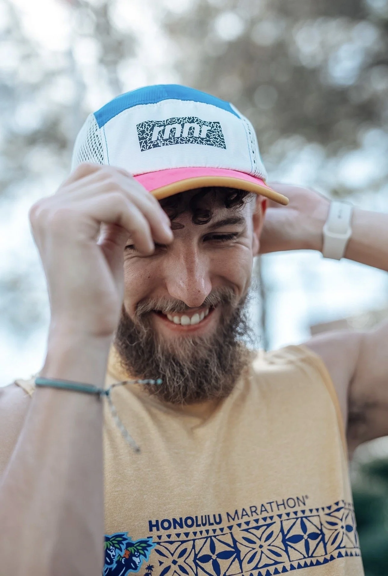 A smiling man with curly hair and a beard adjusting his cap, wearing a beige sleevelessshirt with Honolulu Marathon printed on it, outdoors with blurred background.