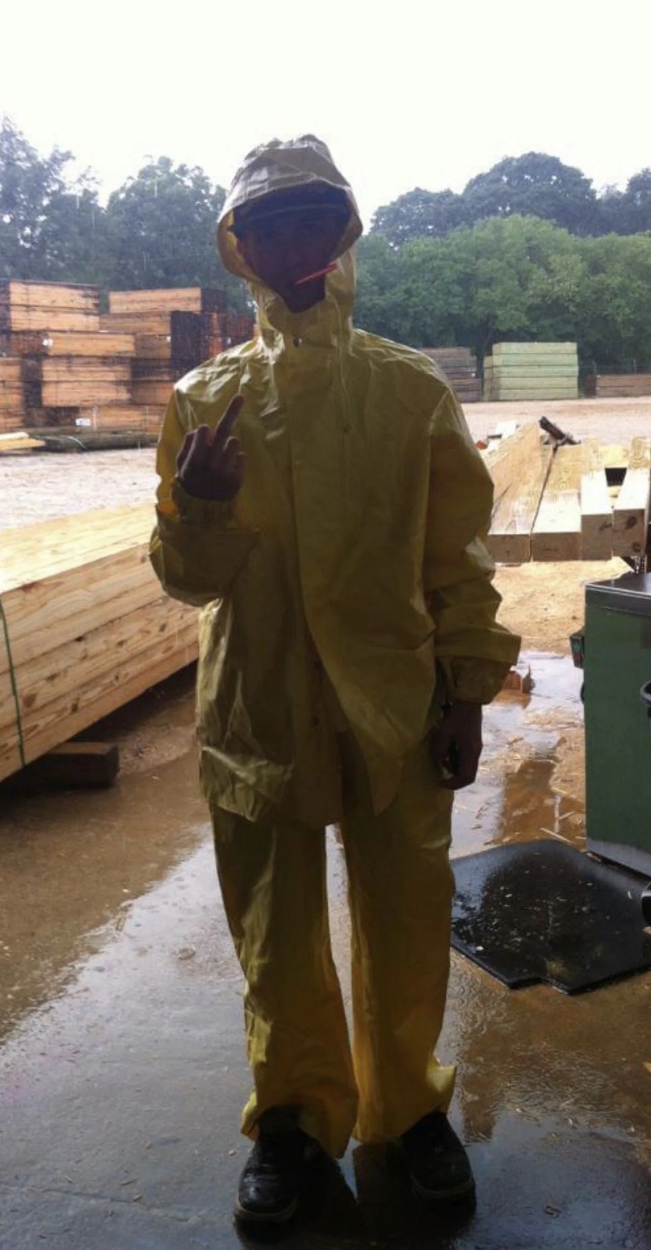 Person wearing yellow rain gear standing in a woodworking yard, holding up middle finger, with stacks of lumber in background.