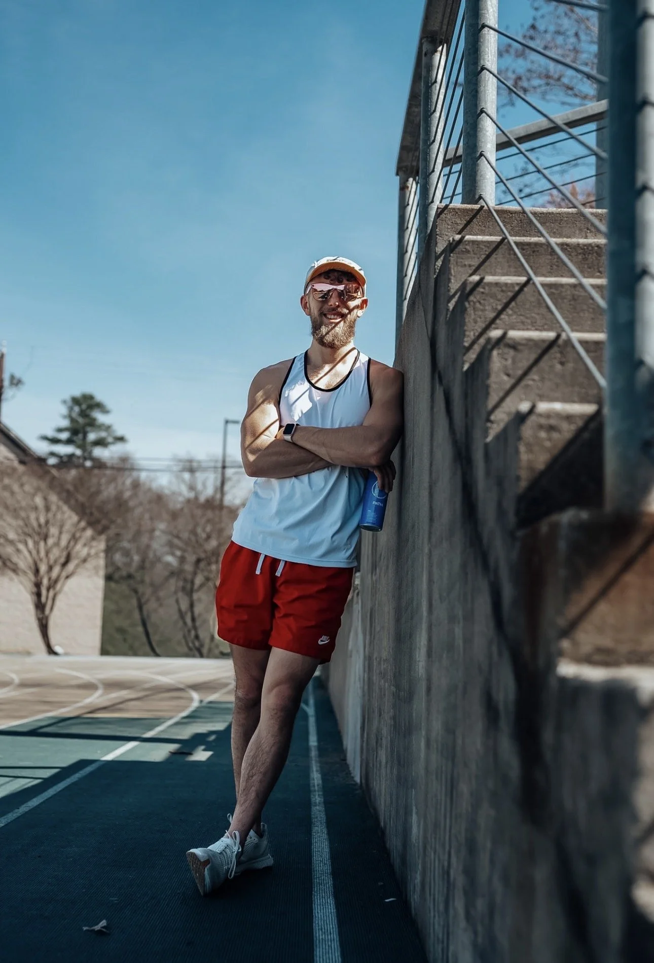 A man wearing a white tank top, red shorts, and sunglasses leaning against a wall on a running track, holding a water bottle, smiling on a sunny day.