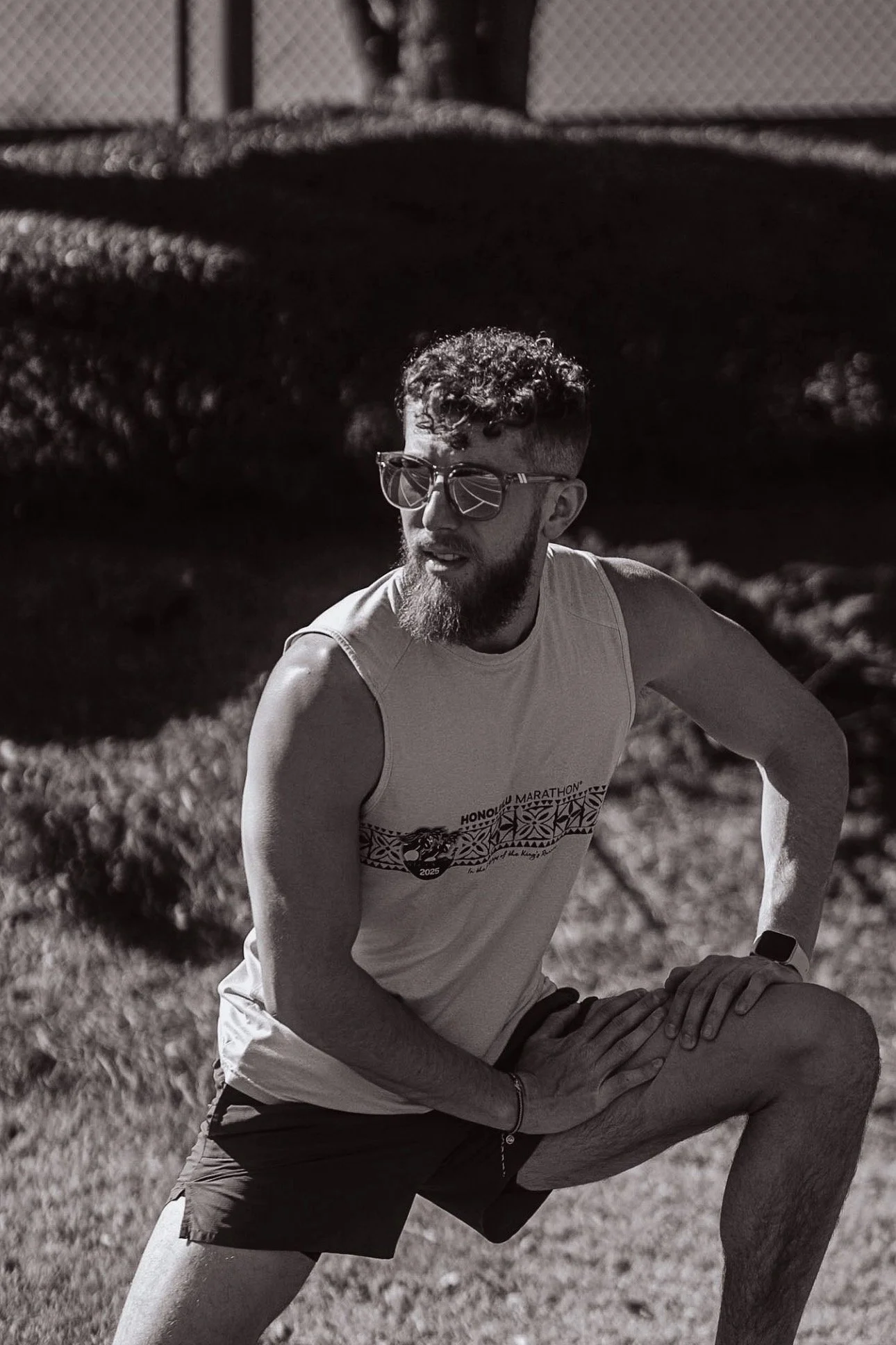 A man with curly hair, beard, and sunglasses crouches on a grassy field, wearing a sleeveless shirt and shorts, with a fence in the background.