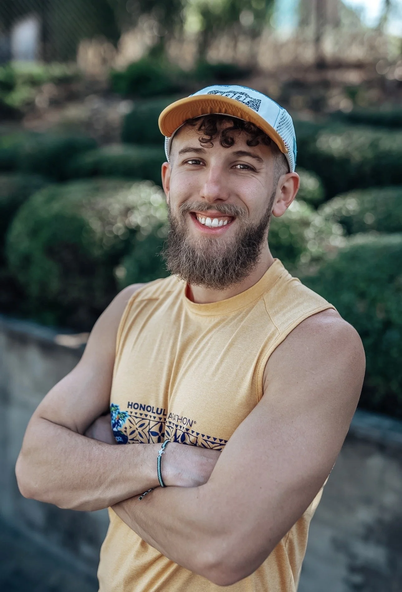 Young man with a beard smiling and standing outdoors with arms crossed, wearing a yellow tank top and a baseball cap, in a natural setting with greenery in the background.