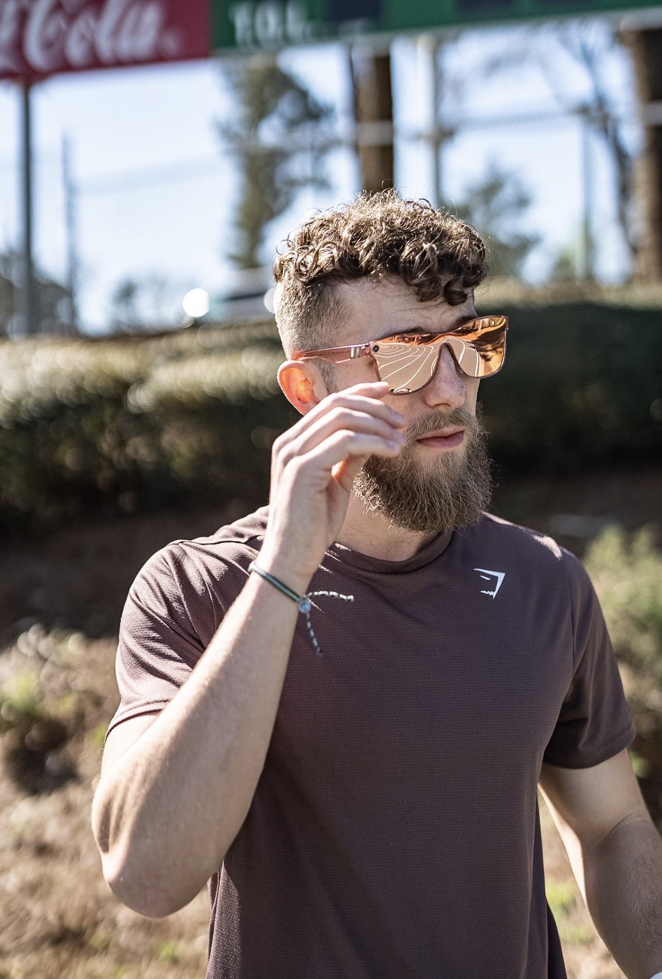 A man with curly hair and a beard wearing reflective sunglasses and a dark athletic shirt outdoors