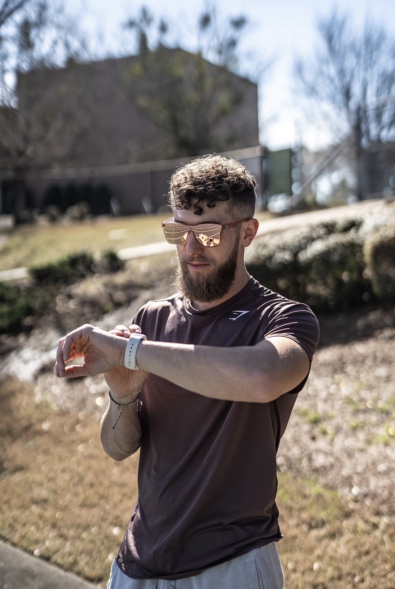 A man with curly hair, sunglasses, and a beard checks his smartwatch outdoors on a sunny day, wearing a brown athletic shirt and gray shorts, with trees and a house in the background.