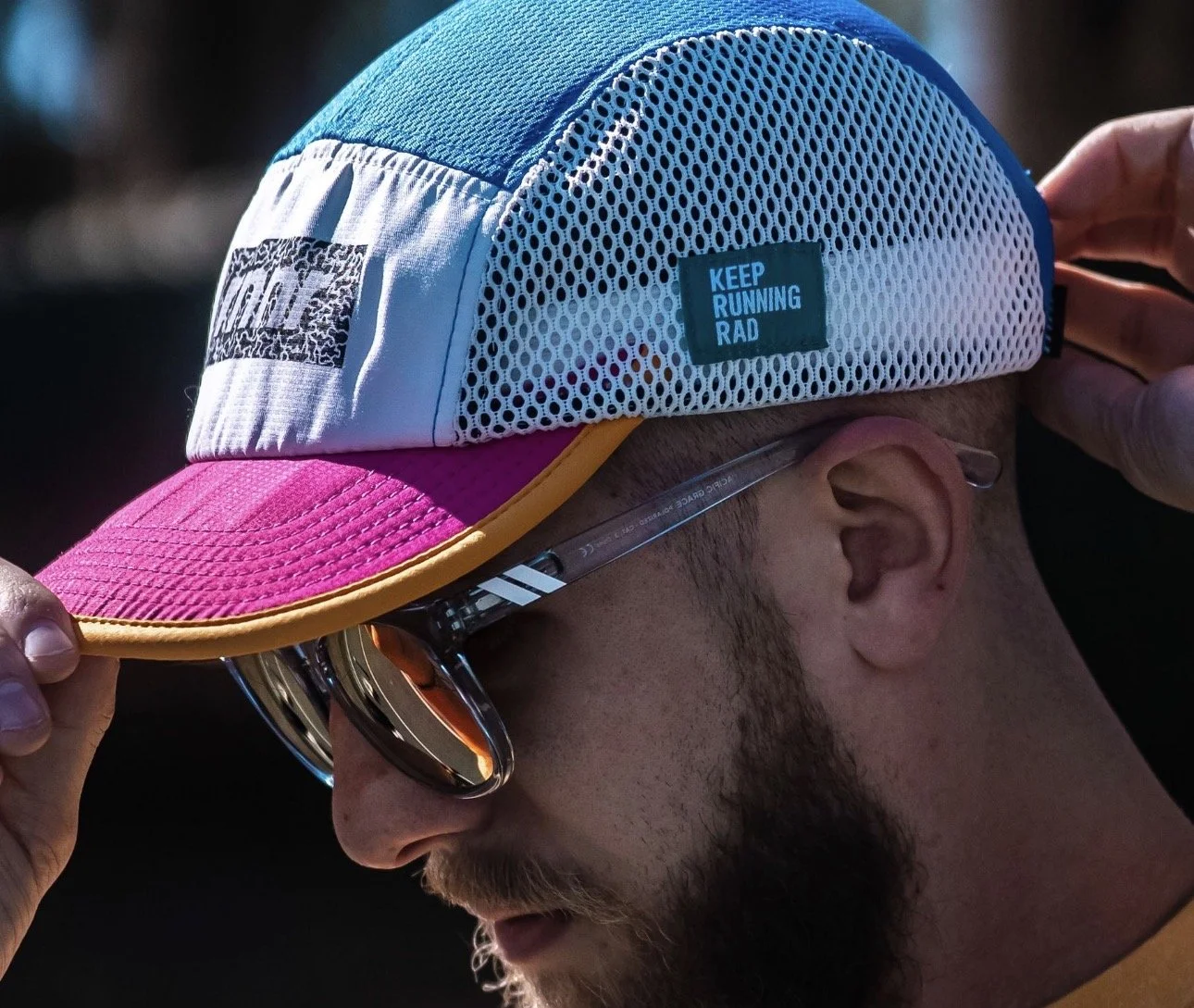 A man wearing a multi-colored running cap and sunglasses adjusting the cap's brim.