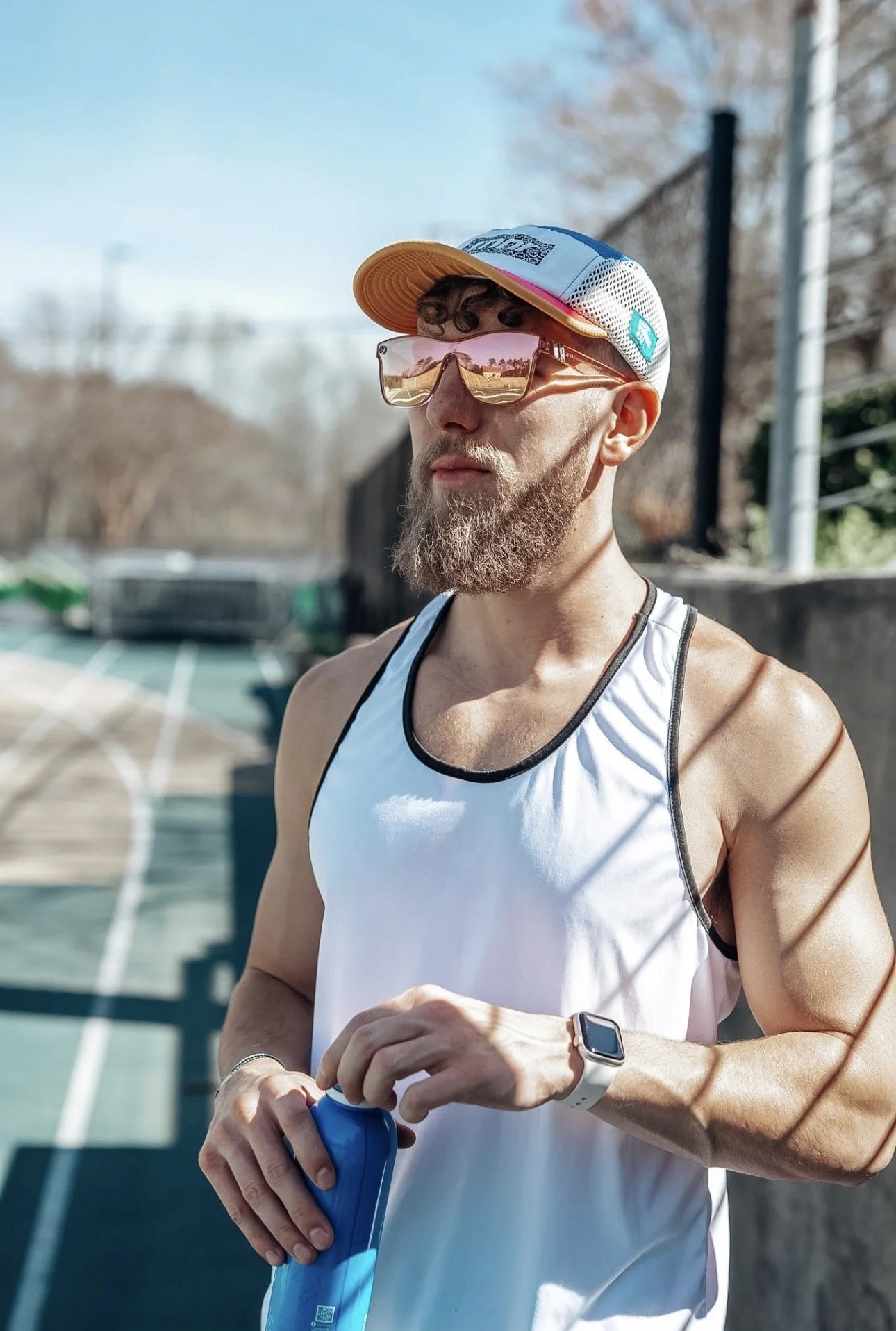 A man with a beard and curly hair wearing sunglasses, a running tank top, a cap, and a smartwatch, standing outdoors on a running track during daytime, holding a blue water bottle.