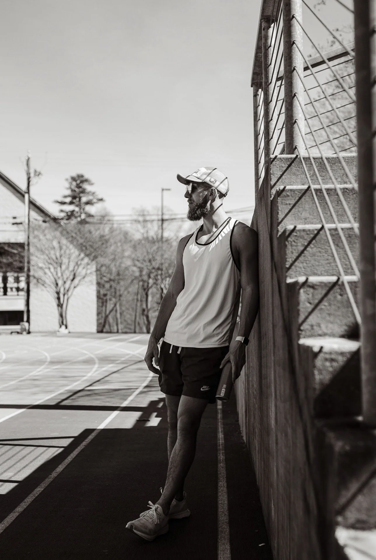 A man with a beard wearing a cap, sunglasses, a sleeveless shirt, shorts, and sneakers standing against a wooden fence on an outdoor running track, holding a water bottle.