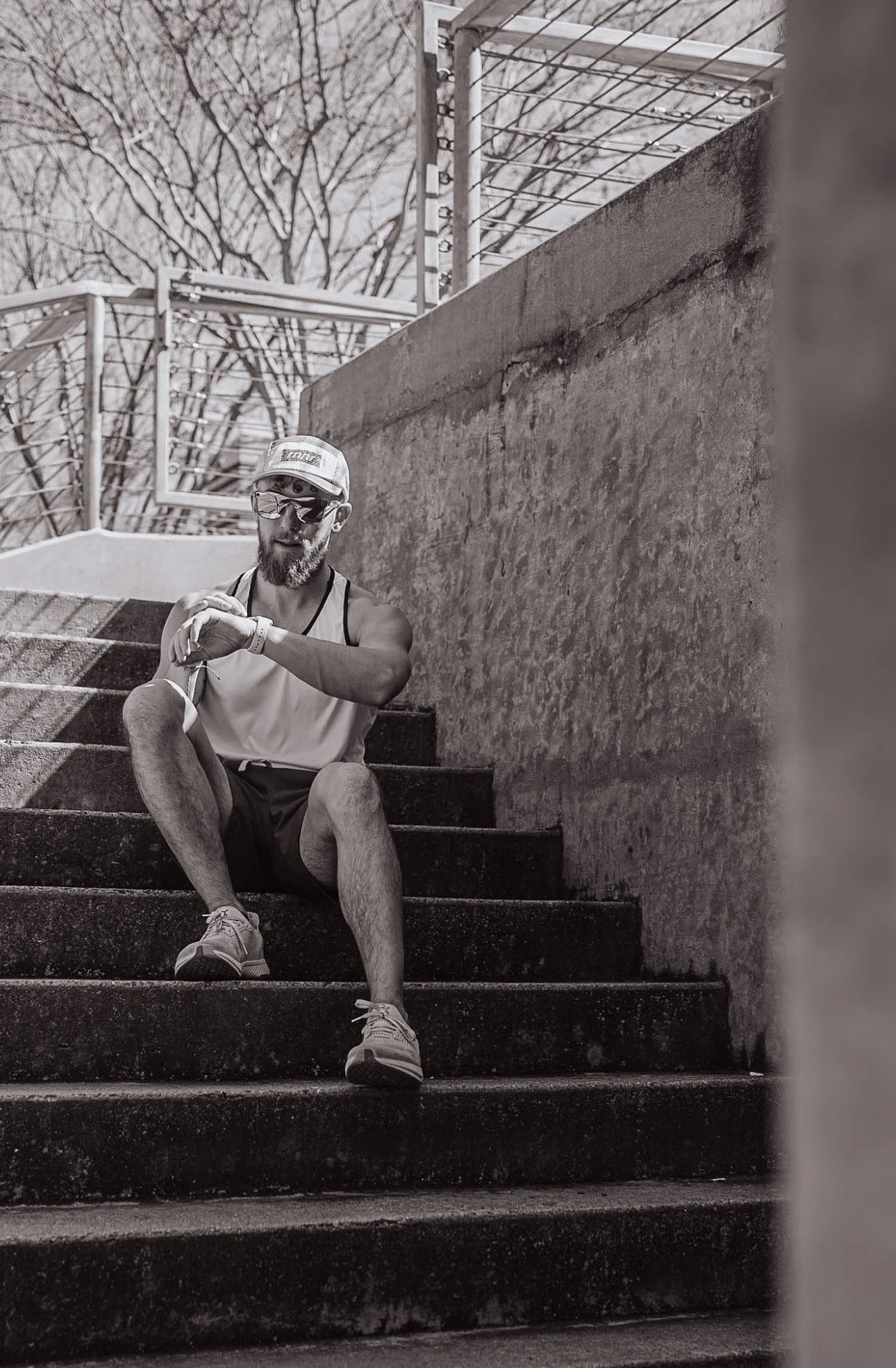 A man sitting on outdoor stairs, checking his watch, wearing sunglasses, a cap, athletic tank top, shorts, and running shoes, with a textured wall on one side and leafless trees in the background.