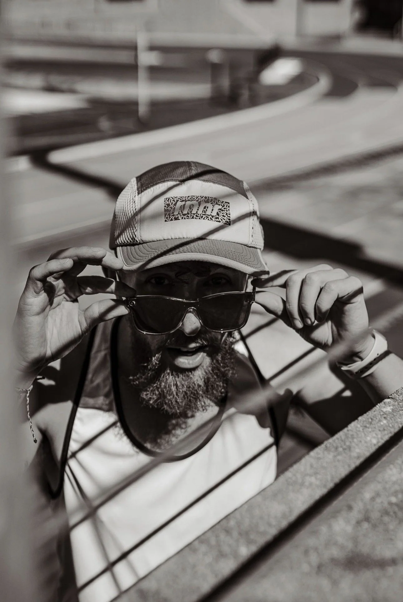 A man with a beard wearing a baseball cap and sunglasses, peeks over a metal railing, holding his sunglasses with both hands.