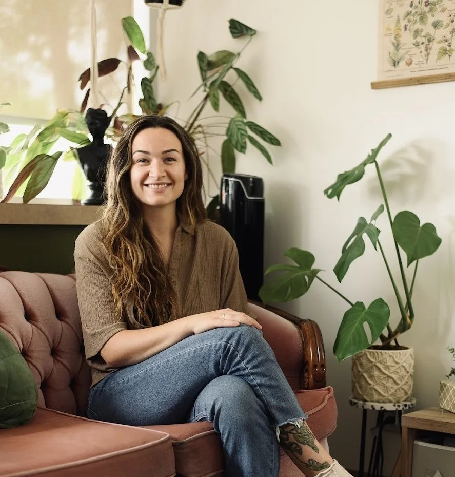 A woman with long wavy hair, smiling, sitting on a pinkish-brown vintage sofa with a green plush pillow. She's wearing a brown shirt and blue jeans, with a tattoo visible on her lower leg. The room has various potted plants, a dress form bust, and botanical prints on the wall, creating a cozy, plant-filled space.