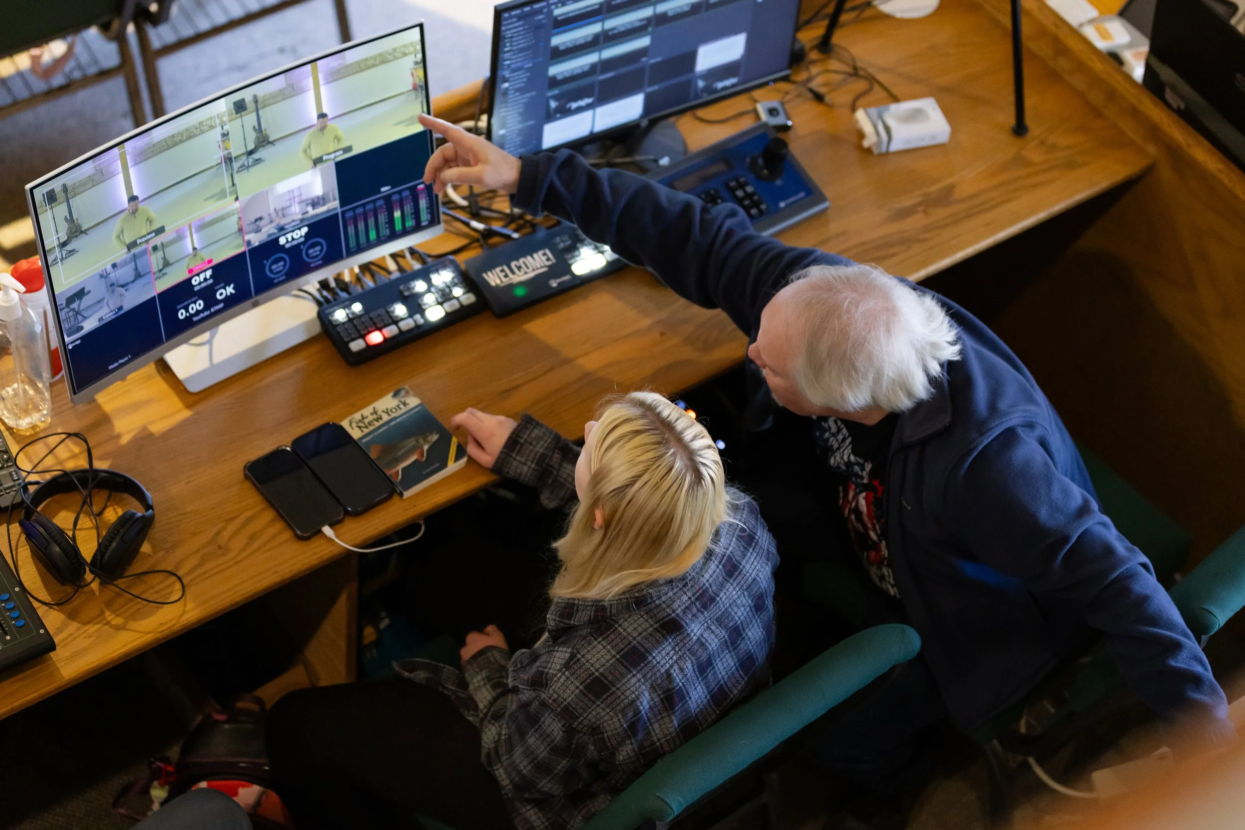 An elderly man with grey hair, sitting with two young children at a wooden table, pointing at a large monitor displaying video editing software. The children are looking at the screen, surrounded by headphones, smartphones, and a book titled 'Fins & Tails of New York'.