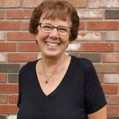 A smiling woman with short brown hair, glasses, and a black shirt, standing in front of a brick wall.