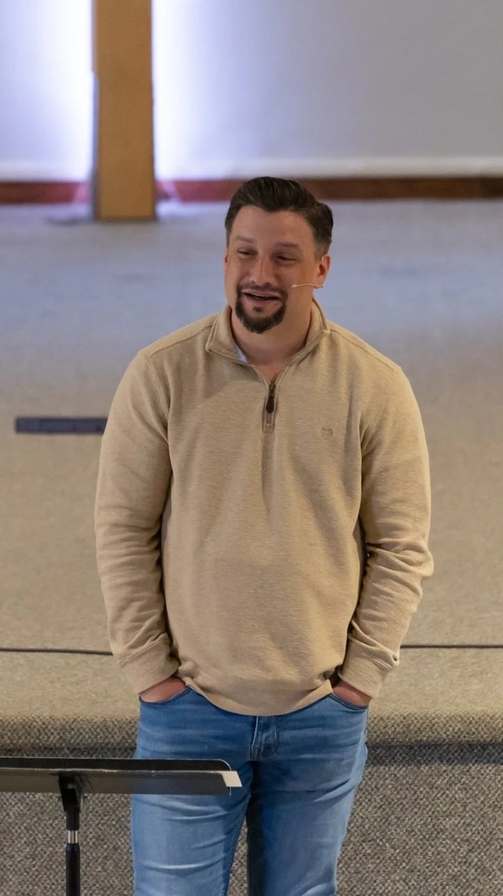 A man with dark hair and a goatee, wearing a beige zip-up sweater and blue jeans, standing with his hands in his pockets and smiling inside a room with wooden and carpeted floors.