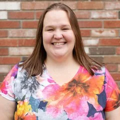 Smiling woman with shoulder-length brown hair wearing a colorful floral shirt, standing in front of a brick wall.