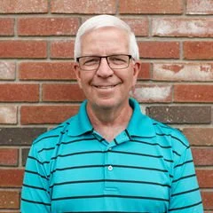 A smiling older man with white hair and glasses, wearing a teal striped polo shirt, standing in front of a brick wall.