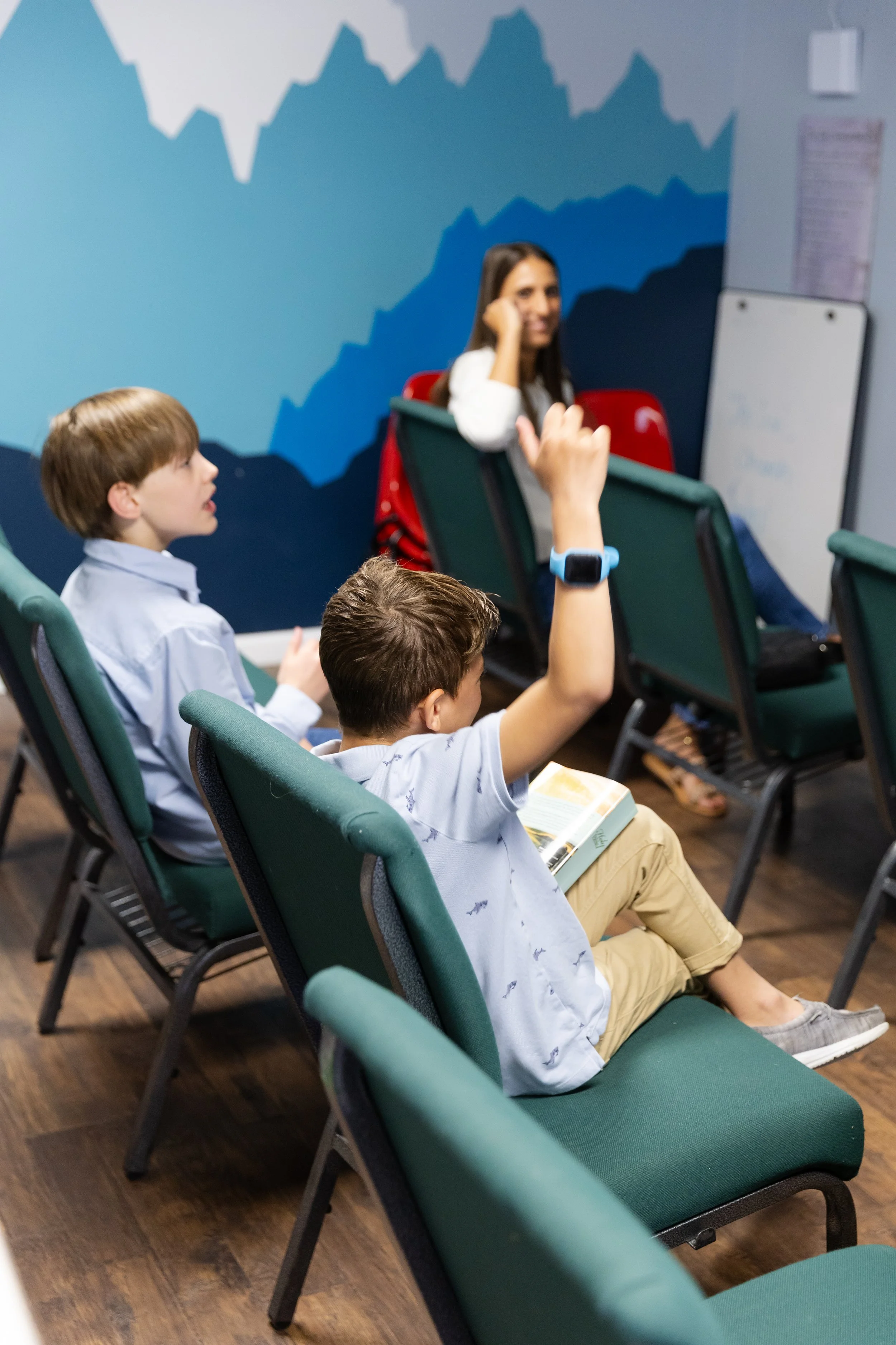 Young students in classroom, one raising hand, teacher listening, mountain chart painted on wall, students seated in green chairs, wood floor, classroom setting.