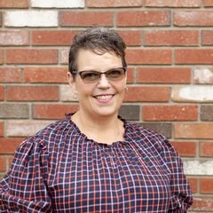 Woman wearing glasses and a checkered shirt, smiling, standing in front of a brick wall.