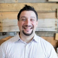 A smiling man with dark hair and a goatee, wearing a light-colored checkered shirt, standing in front of a wooden wall background.
