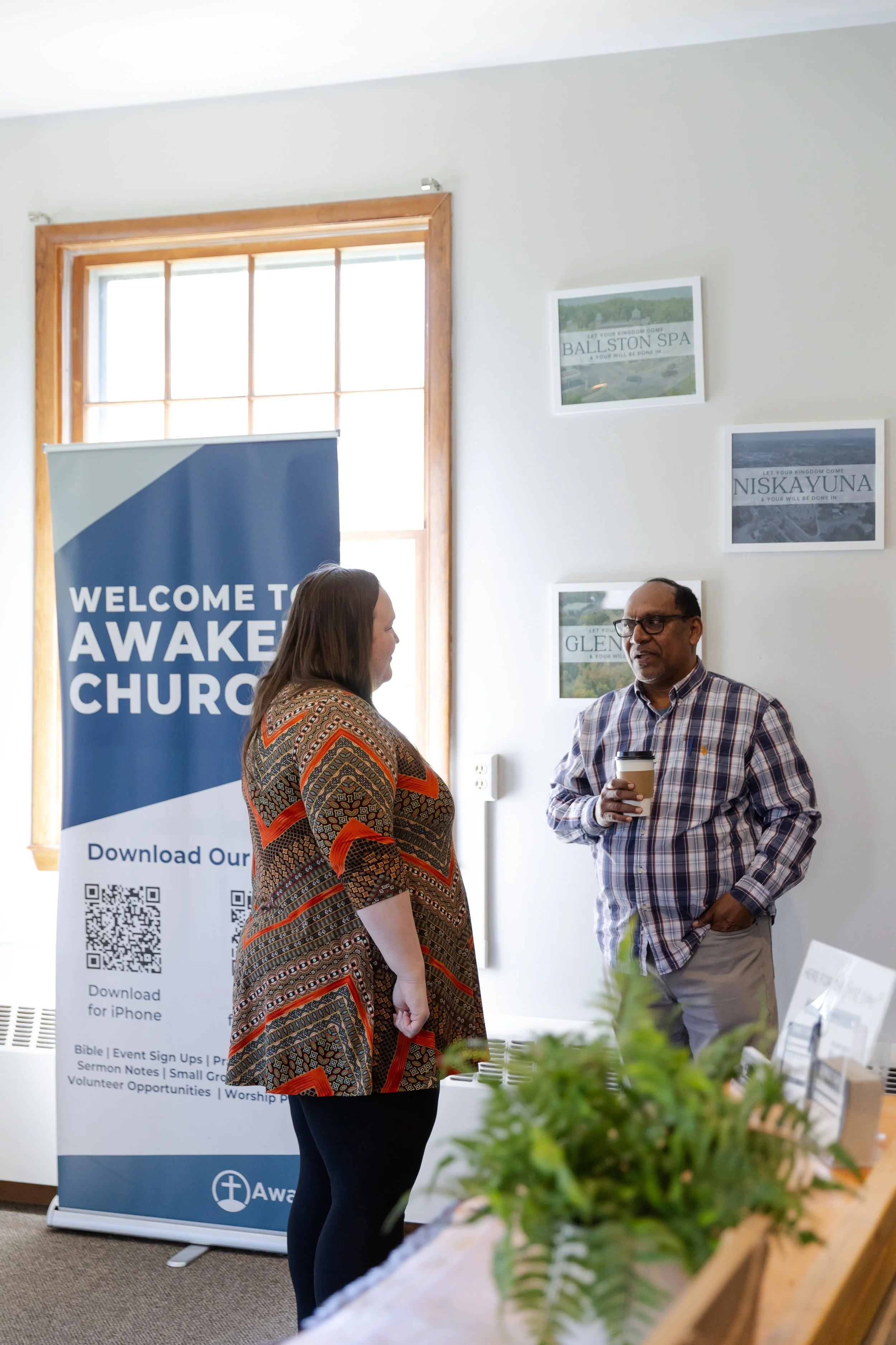 Two people conversing indoors, one woman and one man, near a large window with a church banner and framed pictures on the wall. The woman has long hair and wears a patterned dress, and the man has glasses and wears a plaid shirt, holding a coffee cup. A plant is partially visible in the foreground.