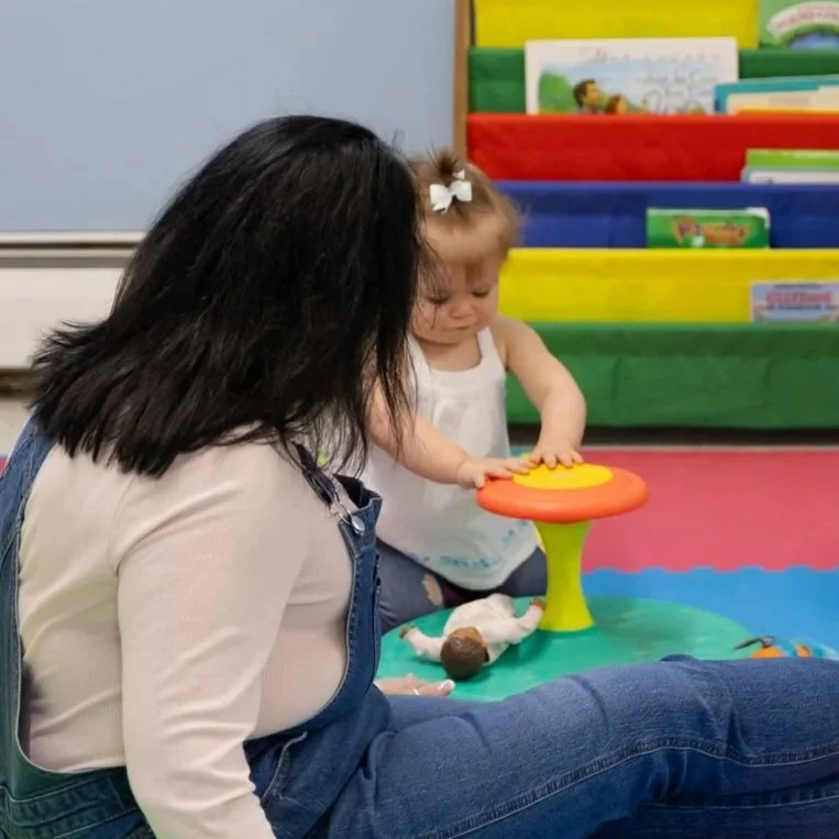 A woman and a young girl sitting on the floor in a classroom or play area, playing with a colorful toy.