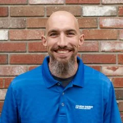 A man with a shaved head and beard smiling, wearing a blue polo shirt, standing in front of a brick wall.