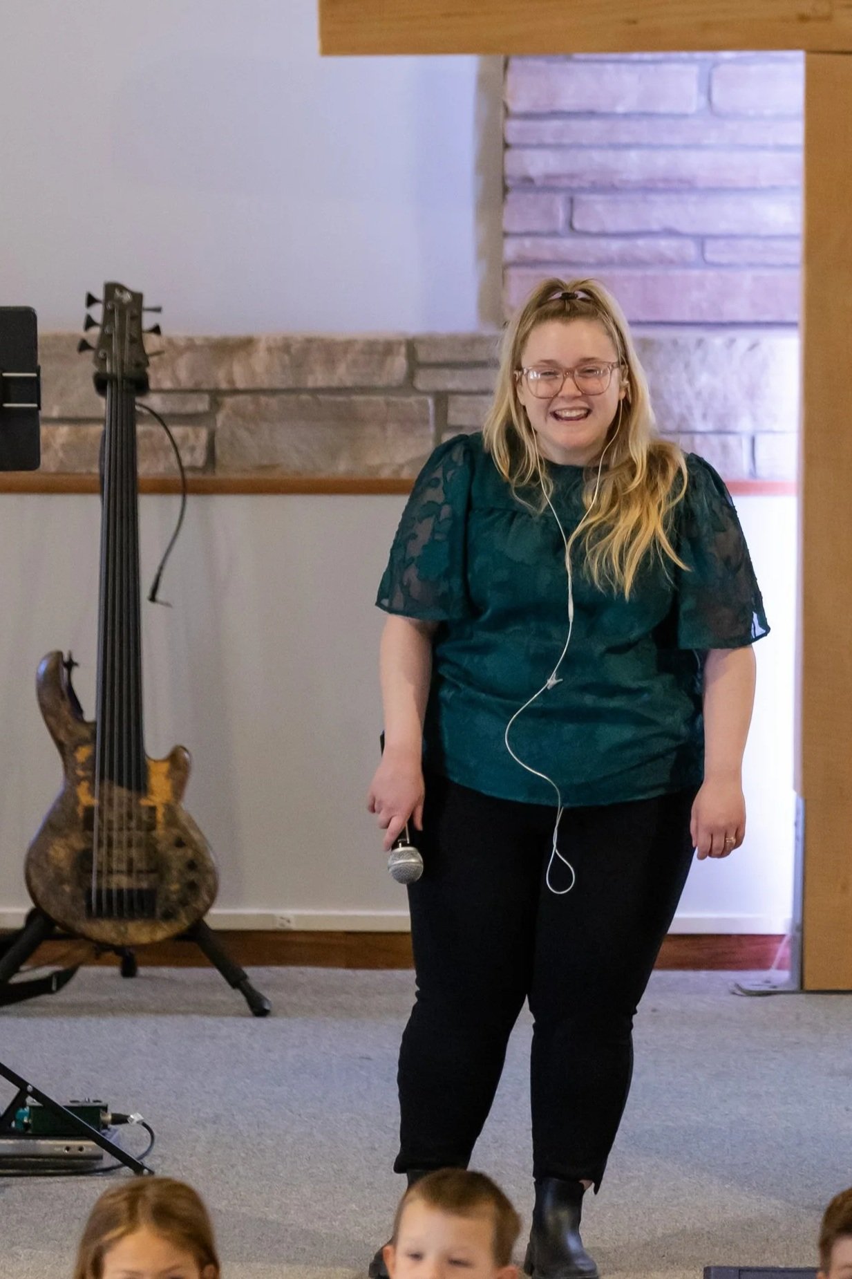 A woman with blonde hair, glasses, wearing a dark green top and black pants, smiling and holding a microphone, standing in front of a group of children in an indoor setting.