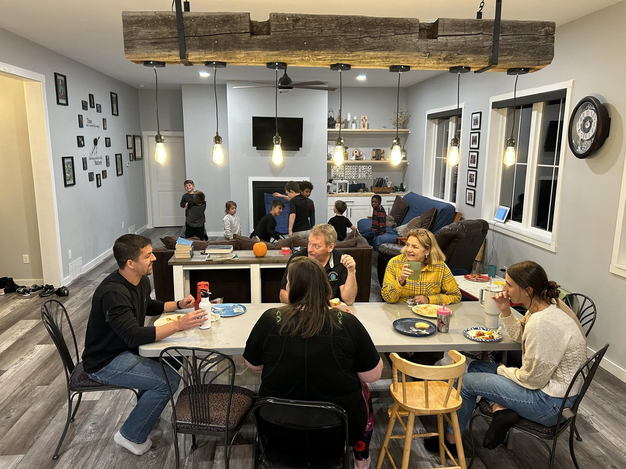 A family gathering in a living room with children and adults seated at a dining table and sofa, enjoying a meal and conversation, with a rustic wooden beam ceiling and framed pictures on the walls.